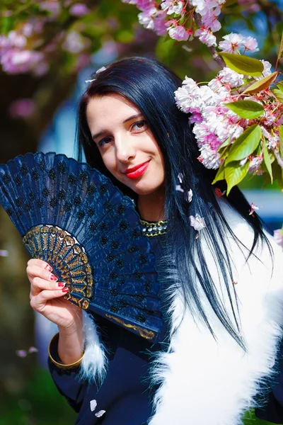 Beautiful Girl with flowers, glamour white fur and black fan in hand, posing next to blooming magical spring rosa sakura flowers. Flower background — Stock Photo, Image