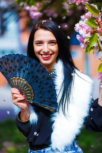 Hermosa chica con flores, glamour de piel blanca y abanico negro en la mano, posando junto a flores mágicas de primavera rosa sakura. Fondo de flores — Foto de Stock