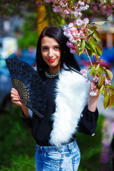 Beautiful Girl with flowers, glamour white fur and black fan in hand, posing next to blooming magical spring rosa sakura flowers. Flower background — Stock Photo, Image