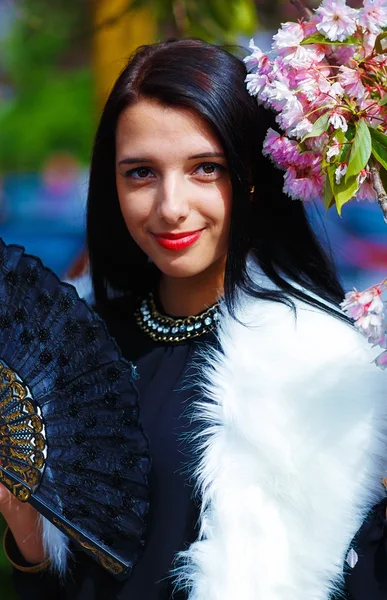 Beautiful Woman with flowers, glamour white fur and black fan in hand, posing next to blooming magical spring rosa sakura flowers. Flower background — Stock Photo, Image