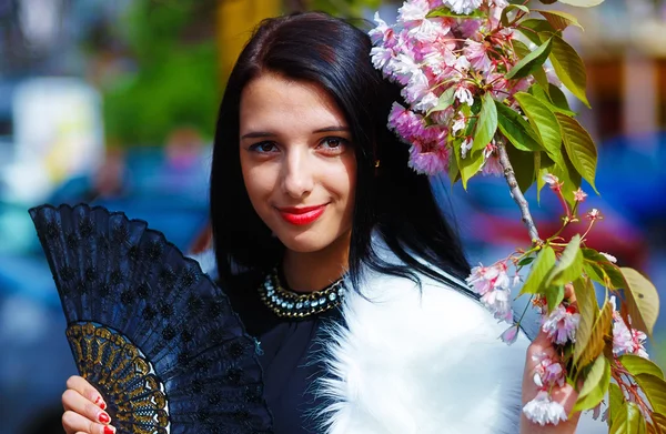 Девушка с цветами, гламурным белым мехом и черным веером в руках, позирующая рядом с цветущей волшебной весной и цветами сакуры. Цветочный фон — стоковое фото