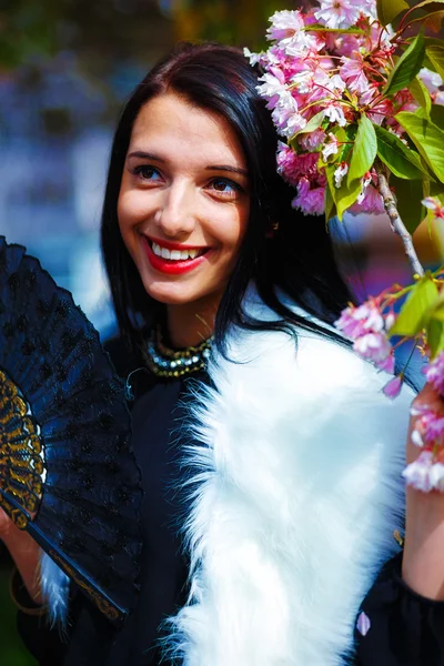 Hermosa chica con flores, glamour de piel blanca y abanico negro en la mano, posando junto a flores mágicas de primavera rosa sakura. Fondo de flores — Foto de Stock