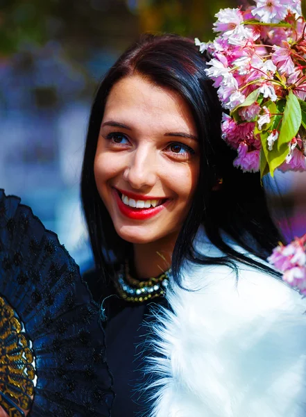 Beautiful Woman with flowers, glamour white fur and black fan in hand, posing next to blooming magical spring rosa sakura flowers. Flower background — Stock Photo, Image