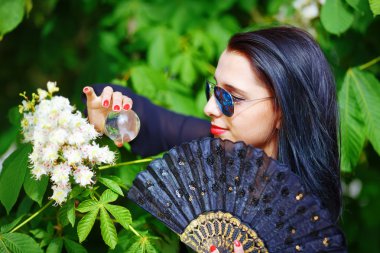 Young woman smelling a bYoung woman smelling a beautiful sakura blossom, purple flowers. Spring Magic, Magical Glass sphere in hand and beautiful ornamental fan. Flower background.