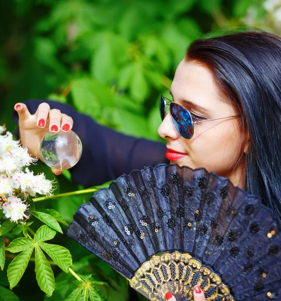 Mujer joven oliendo una bMujer joven oliendo una hermosa flor de sakura, flores púrpuras. Spring Magic, esfera de vidrio mágico en la mano y hermoso abanico ornamental. Fondo de flores . — Foto de Stock