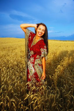 Gülümseyen gün batımı ile buğday alan üzerinde duran süs elbise ile genç kadın. Doğal arka plan ve mavi gökyüzü.