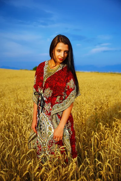 Uśmiechnięta młoda kobieta z ozdobnych sukienka stojący na polu pszenicy z zachodu słońca. Naturalne podłoże i błękitne niebo — Zdjęcie stockowe