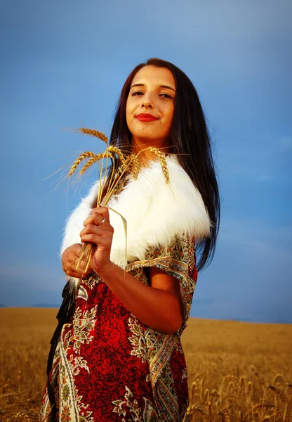 Young woman with ornamental dress and white fur standing on a wheat field with sunset. Natural background — Stockfoto