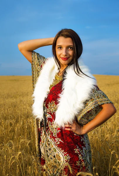 Young woman with ornamental dress and white fur standing on a wheat field with sunset. Natural background — Stock Photo, Image