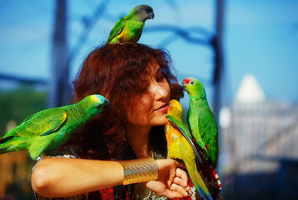 Mulher nova em vestido ornamental e jóias de ouro bonitas com uns papagaios a cores. mulher beijar pássaro . — Fotografia de Stock