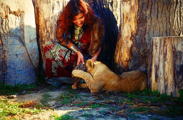 Giovane donna con abito ornamentale e gioiello in oro che gioca con cucciolo di leone in natura . — Foto Stock