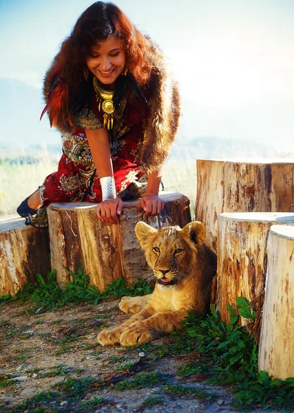 Young woman with ornamental dress and gold jewel playing with lion cub in nature, with wolf fur. — Stock Photo, Image