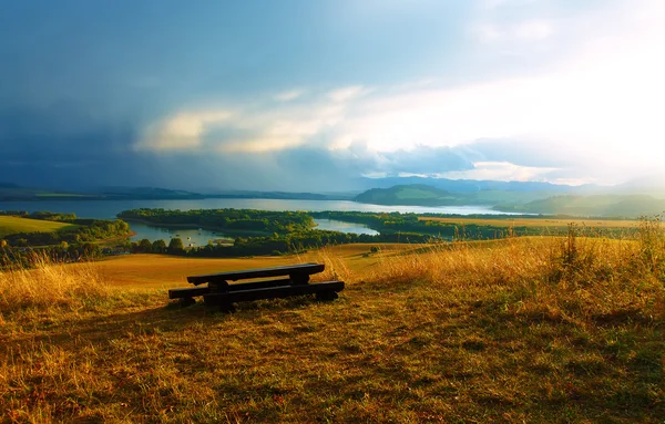 Frumos peisaj. Bancă de lemn pe pajiște, cu vedere la lac și munți cu cer frumos înnorat .. — Fotografie, imagine de stoc