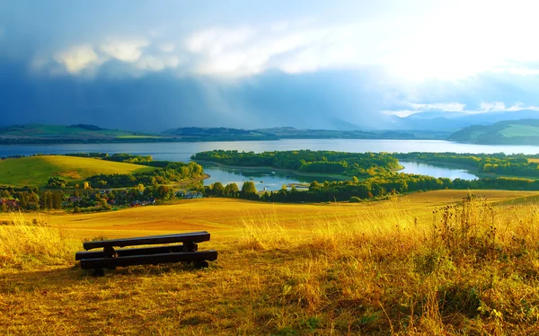 Hermoso paisaje. Banco de madera en el prado, con vistas al lago y las montañas y pueblo con hermoso cielo nublado —  Fotos de Stock