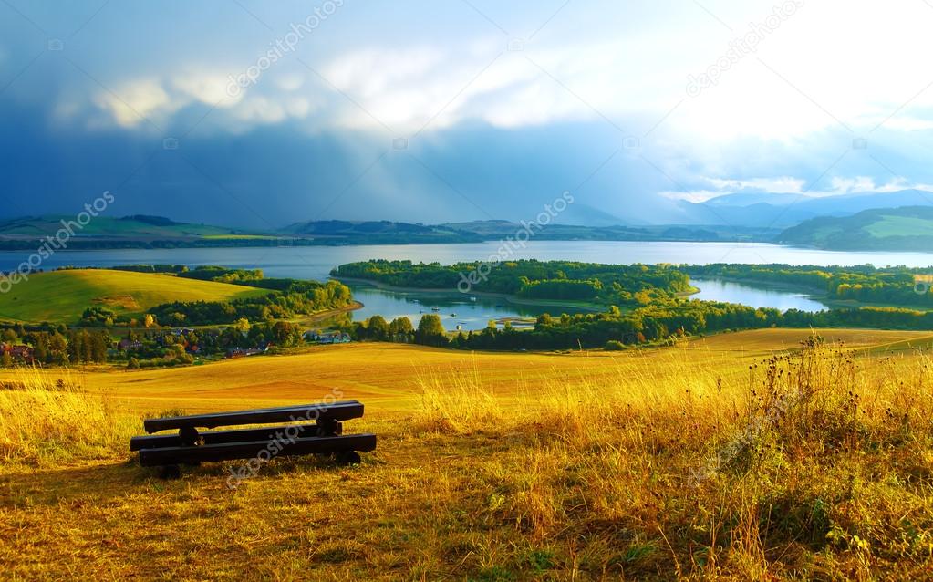 Beautiful landscape. Wooden bench in the meadow, overlooking the lake and mountains  and village with beautiful cloudy sky