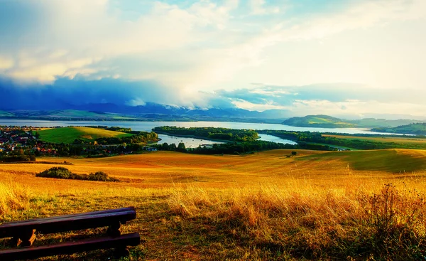 Frumos peisaj. Bancă de lemn pe pajiște, cu vedere la lac și munți și sat cu cer frumos înnorat — Fotografie, imagine de stoc
