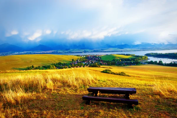 Hermoso paisaje. Banco de madera en el prado, con vistas al lago y las montañas y pueblo con hermoso cielo nublado —  Fotos de Stock