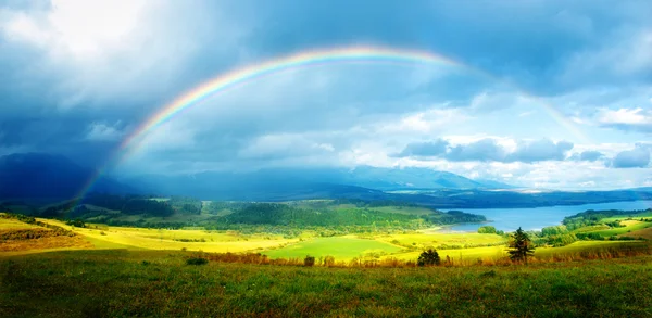 Hermoso paisaje, prado verde y amarillo y lago con la montaña en el fondo con un arco iris en el cielo —  Fotos de Stock