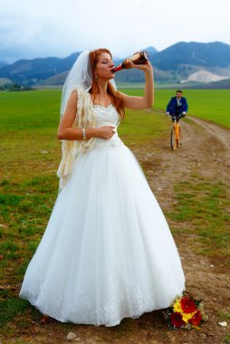 bride with a beer bottle and a groom on bicycle on the background - wedding concept. clipart