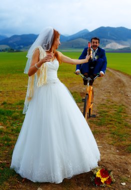 bride with a beer bottle and a groom on bicycle on the background - wedding concept. clipart