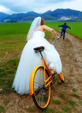 bride on orange retro bike is chasing after a groom in blue wedding suit with a beer bottle. wedding concept. clipart