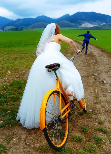Bruid op oranje retro fiets is jagen na een bruidegom in blauwe bruiloft pak met een bierflesje. bruiloft concept. — Stockfoto