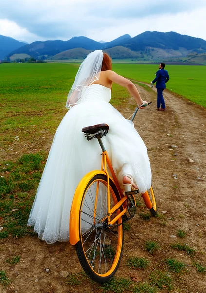 Sposa su arancione bici retrò sta inseguendo dopo uno sposo in abito da sposa blu con una bottiglia di birra. concetto di matrimonio . — Foto Stock