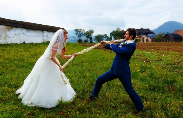 Braut zieht ihren Bräutigam mit Seil zu sich - lustiges Hochzeitskonzept. — Stockfoto