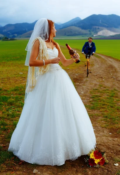 Sposa con una bottiglia di birra e uno sposo in bicicletta sullo sfondo - concetto di matrimonio . — Foto Stock