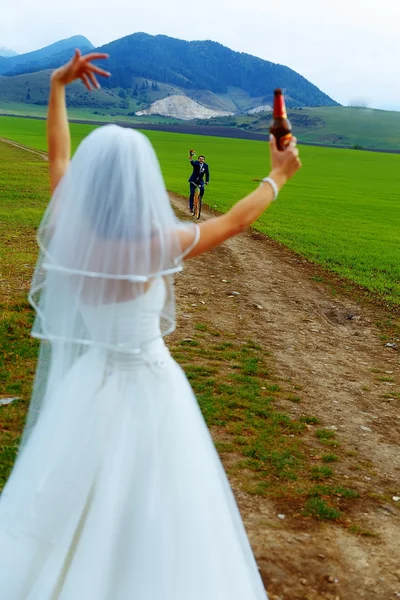 Braut mit Bierflasche und Bräutigam auf Fahrrad im Hintergrund - Hochzeitskonzept. — Stockfoto