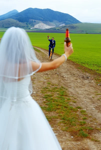 Noiva com uma garrafa de cerveja e um noivo na bicicleta no fundo - conceito de casamento . — Fotografia de Stock