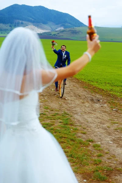 Bruid met een bierfles en een bruidegom op fiets op de achtergrond - bruiloft concept. — Stockfoto