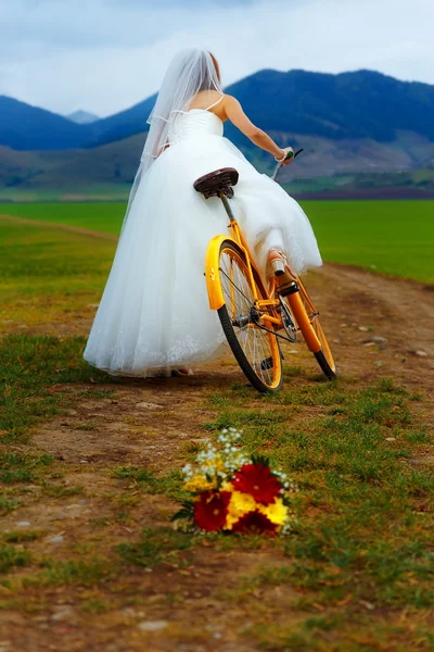 Braut auf orangefarbenem Fahrrad in wunderschönem Brautkleid mit Spitze in der Landschaft. mit Brautstrauß. Hochzeitskonzept. — Stockfoto