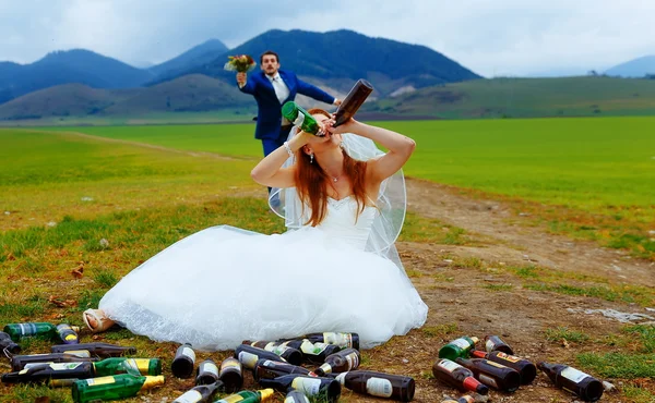 Sposa ubriaca con un sacco di bottiglie di birra vuote nel paesaggio montano e sposo comming a le- concetto di matrimonio divertente . — Foto Stock