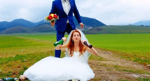 Noiva bêbada com muitas garrafas de cerveja vazias na paisagem da montanha e noivo comemorando com ela conceito de casamento engraçado . — Fotografia de Stock