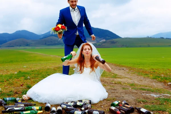 Sposa ubriaca con un sacco di bottiglie di birra vuote nel paesaggio montano e sposo comming a le- concetto di matrimonio divertente . — Foto Stock