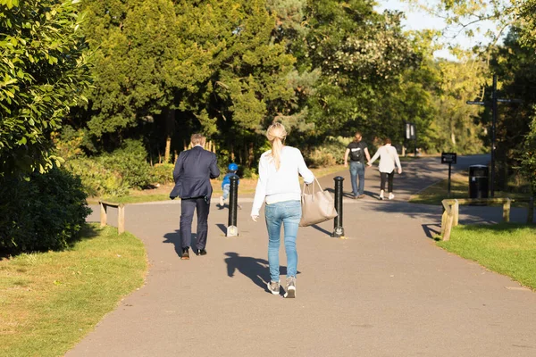 Different Passers-by Walk Along the Asphalt Path of Street Among Green Trees on a Sunny Day. English Park. City People Style, Park Area, Early Autumn. Healthy Walking Every Day.