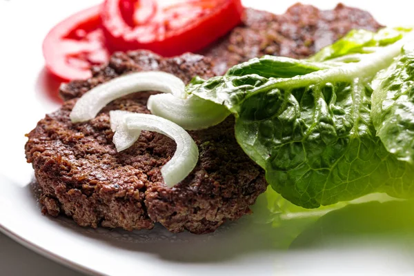 ingredients for a hamburger, looking onto a burger patty with onions, tomato slices and a salad leave on a white plate, to make your own fast food hamburger recipe