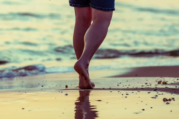 Low angle shot of a woman legs walking at the beach, sunset color theme. Walking with naked feed in the wet sand in summer time.