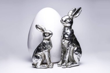Beyaz seramik yumurta önünde iki gümüş eş Paskalya tavşanı figürü, yaratıcı Paskalya ve bahar dekorasyonu 2020, beyaz izole arka planda moda renkleri