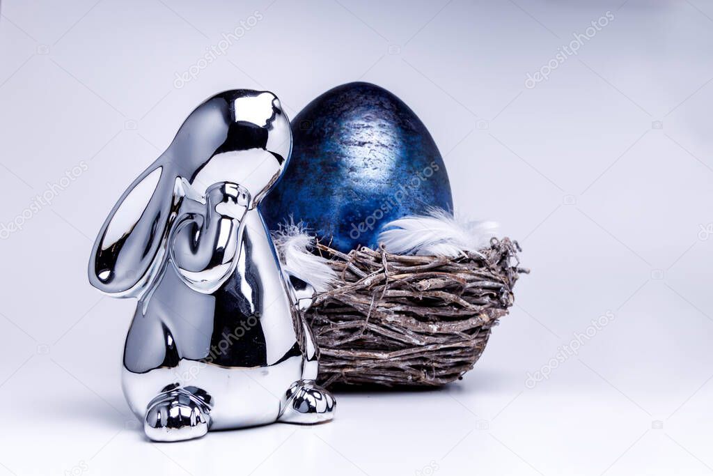 Cute silver metallic easter bunny figure looks onto a royal blue easter egg laying in a easter nest made of brown branches, filled with white feathers on white uniform background