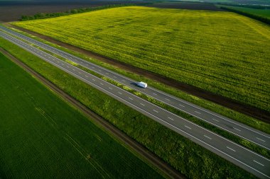 Yeşil tarlalar boyunca asfalt yolda ilerleyen beyaz minibüs. Havadan görüldü. Hava manzaralı. İHA fotoğrafçılığı. seyahat kavramı