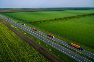 kargo teslimatı ve ulaşım. Yeşil tarlalar boyunca asfalt yolda giden servis kamyonları. Havadan görüldü. Hava manzaralı. İHA fotoğrafçılığı. kargo teslimatı ve ulaşım kavramı sol taraf trafiği