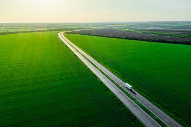 Günbatımında yalnız beyaz bir kamyon. Yük teslimatı asfalt yolda yeşil tarlalar arasında mallarla birlikte gidiyor. Havadan görüldü. Hava manzaralı. Drone fotoğrafçılığı. 