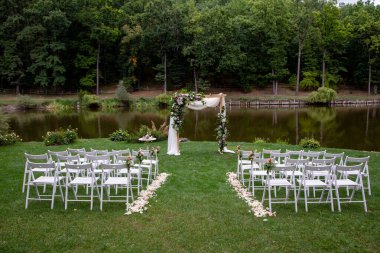 Ormandaki gölün kenarında düğün töreni. Açık havada yapılacak bir düğün için şık bir dekorasyon. Güzel beyaz mobilyaları olan açık hava düğün töreni. Tahta misafir sandalyeleri..
