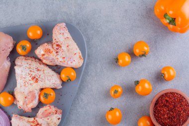 Gri mutfak masasında çiğ tavuk, baharat ve olgun domates.