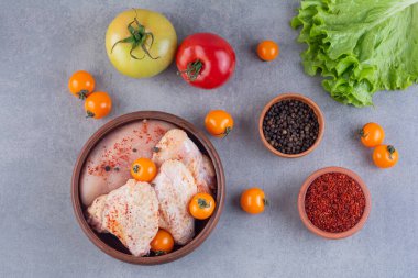 Gri mutfak masasında çiğ tavuk, baharat ve olgun domates.