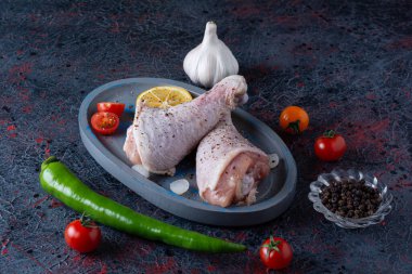 Gri mutfak masasında çiğ tavuk, baharat ve olgun domates.