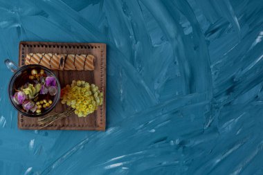 Mavi arka planda kurutulmuş çiçekler ve kurabiyelerle bir fincan çay.