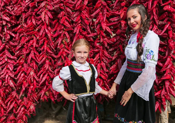 Meninas vestidas com roupas tradicionais sérvias balcânicas, traje popular nacional. Posando perto do lote pimentas de páprica vermelhas. — Fotografia de Stock